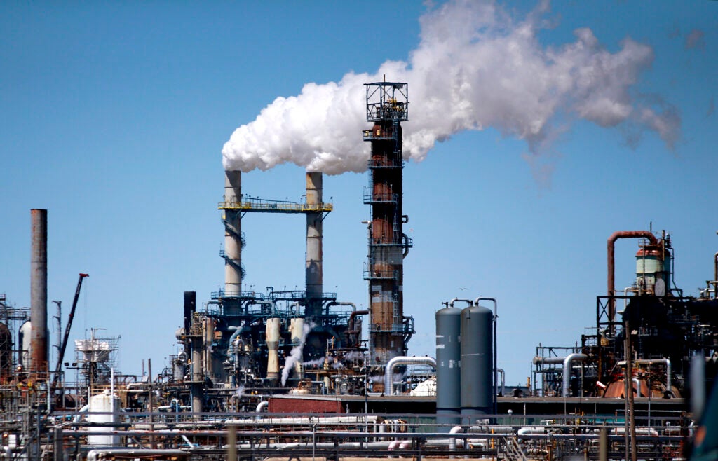 Two smoke stacks emit a large cloud of white smoke into a blue sky, in the middle of a large industrial facility made up of pipes and tanks.
