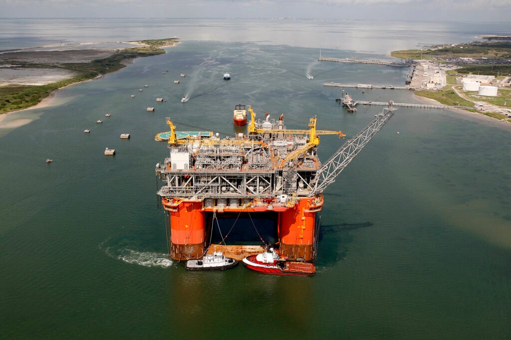 A large red oil platform is surrounded by two tug boats in a channel of water.