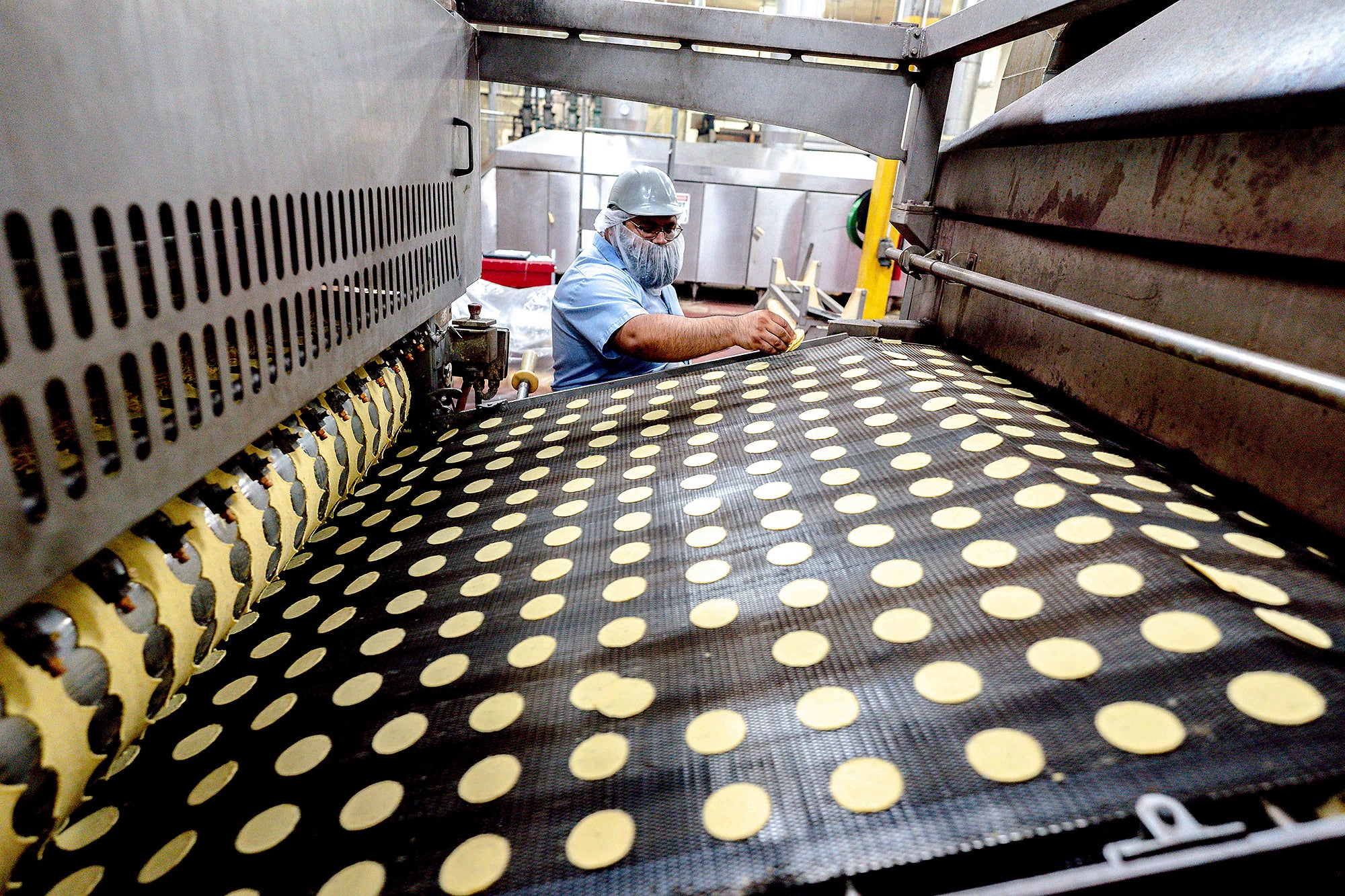 Small round uncooked corn chips move across a conveyer belt as a man wearing a hard hat and beard net inspects them in the background.