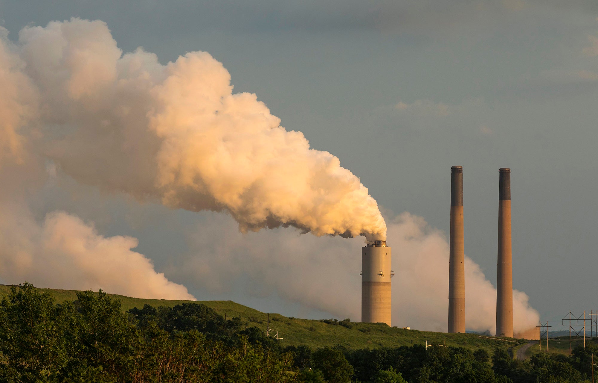 Endangerment Coal-Fired Power Plant Three smokestacks rise from behind a green hill with one spewing a large amount of white smoke. Smoke it also coming out of two other areas obscured by the hill.