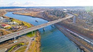 A multilane highway bridge with cars driving on it, above a river. Green fields and buildings on one side of the river. Brown fields to the other. Trees and residential buildings further inland.