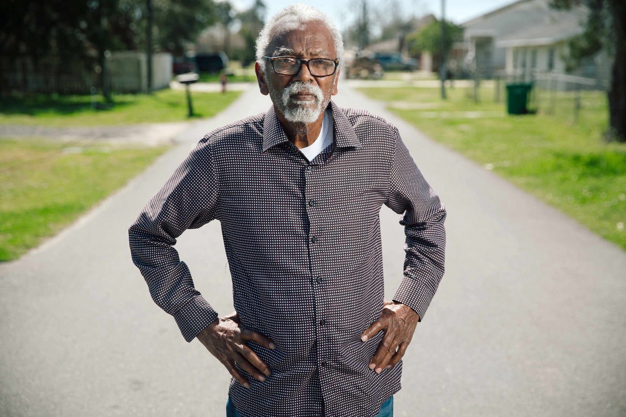 A man with white hair and white facial hair stands in the middle of a residential street. His hands at his hips.