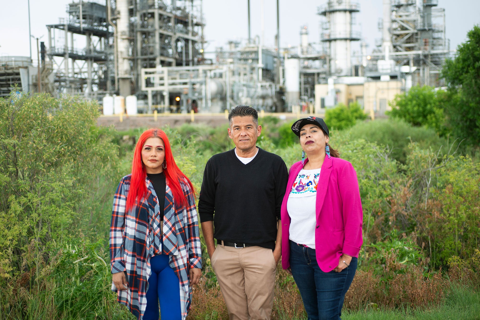 Three people stand amongst vegetation. Background of an oil refinery nearby.