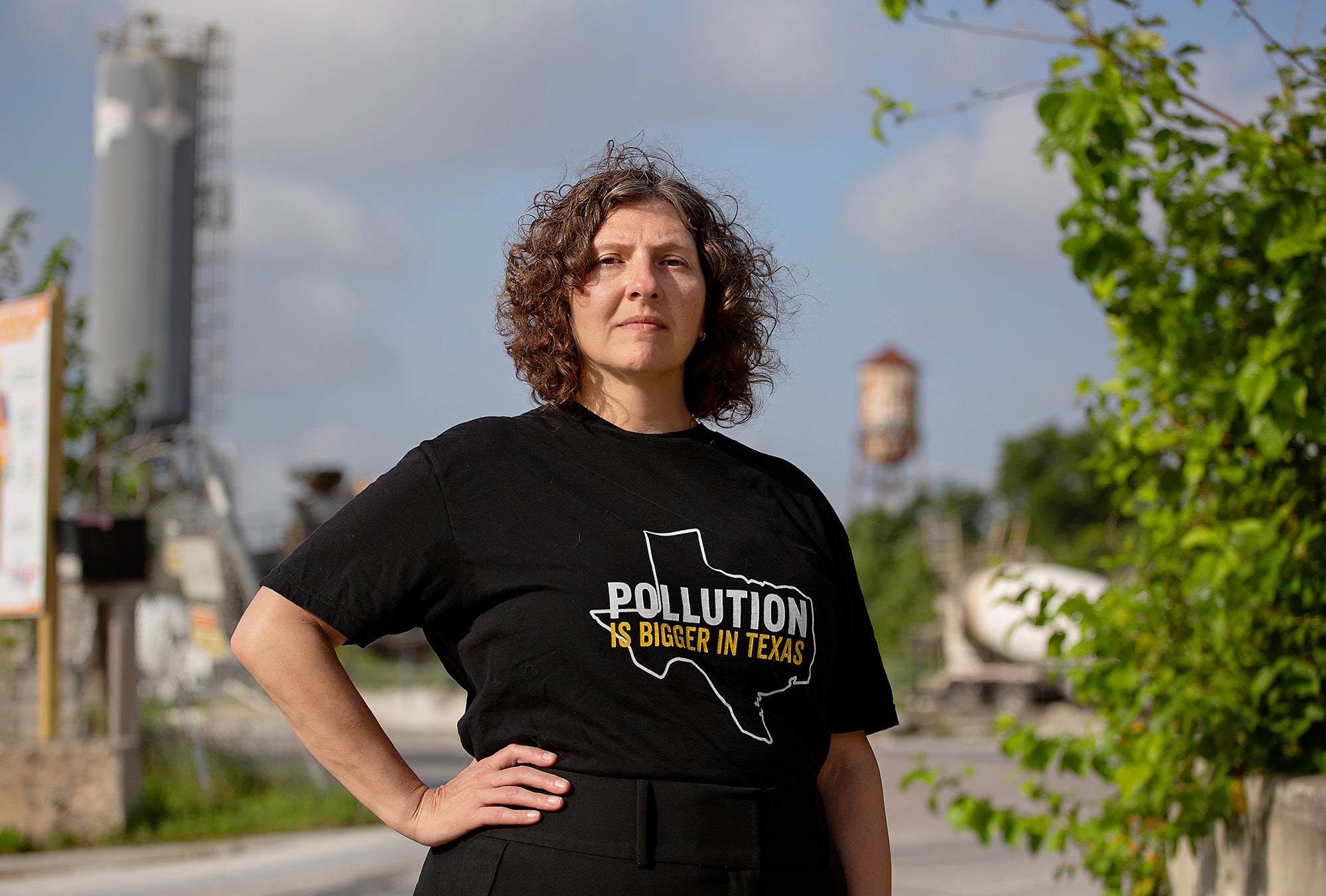 Air Alliance A woman standing next to an industrial facility with a hand on her hip. She is wearing a black t-shirt with an outline of Texas with the words "Pollution is Bigger in Texas"