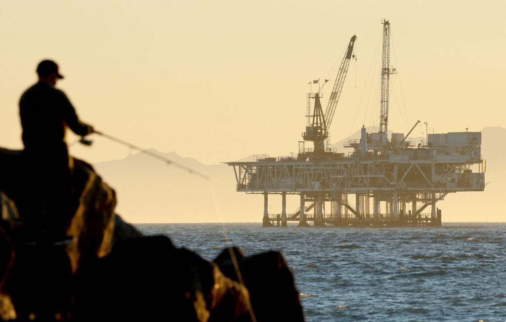 An man fishes on the rocky shore as a large oil platform sits in the water in the distance. It appears to be close to shore.
