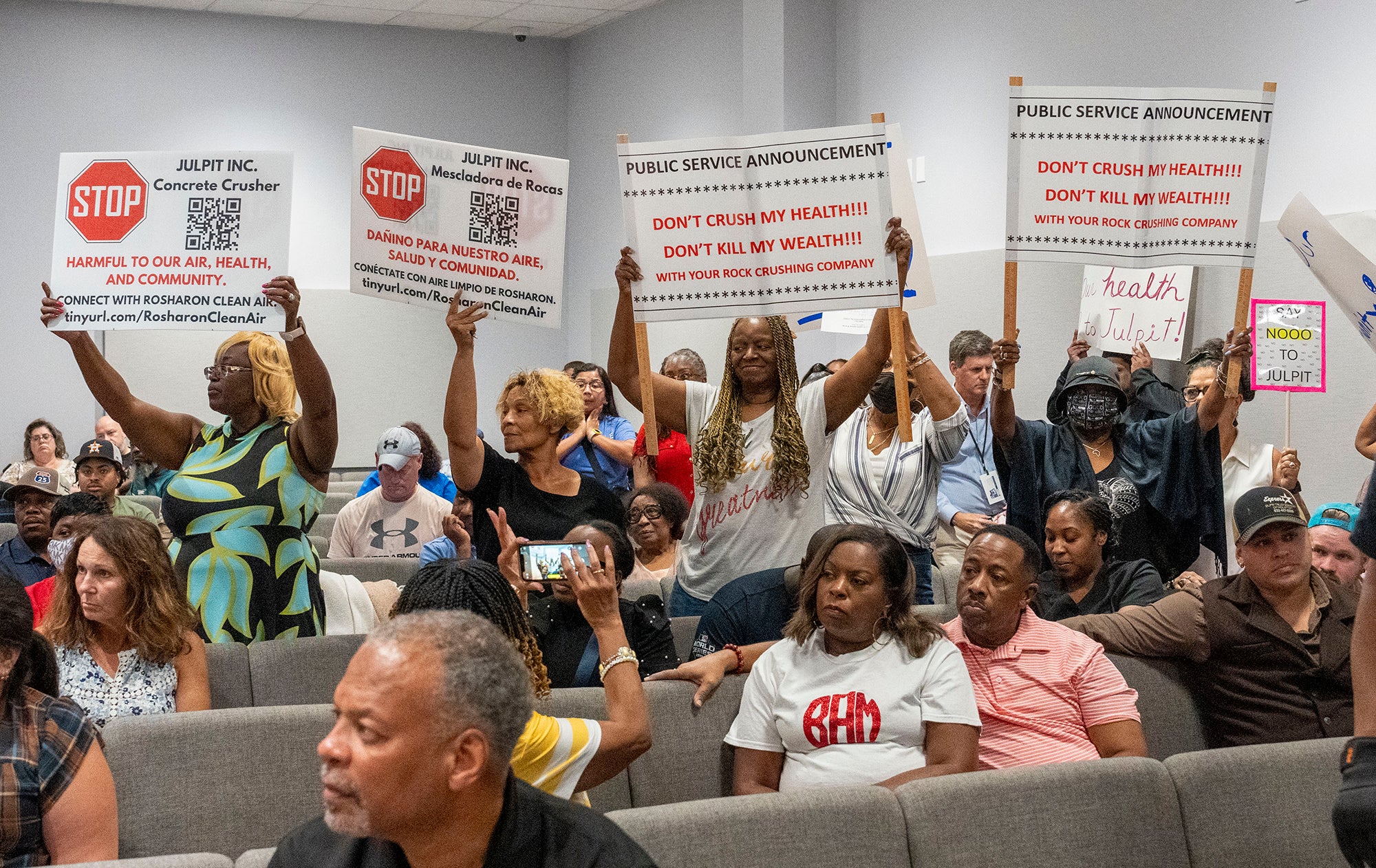 People sit in a meeting room with signs reading "Stop Julpit, Inc." and "Dont' crush my health!!! Don't kill my wealth!!! With your rock crushing company"