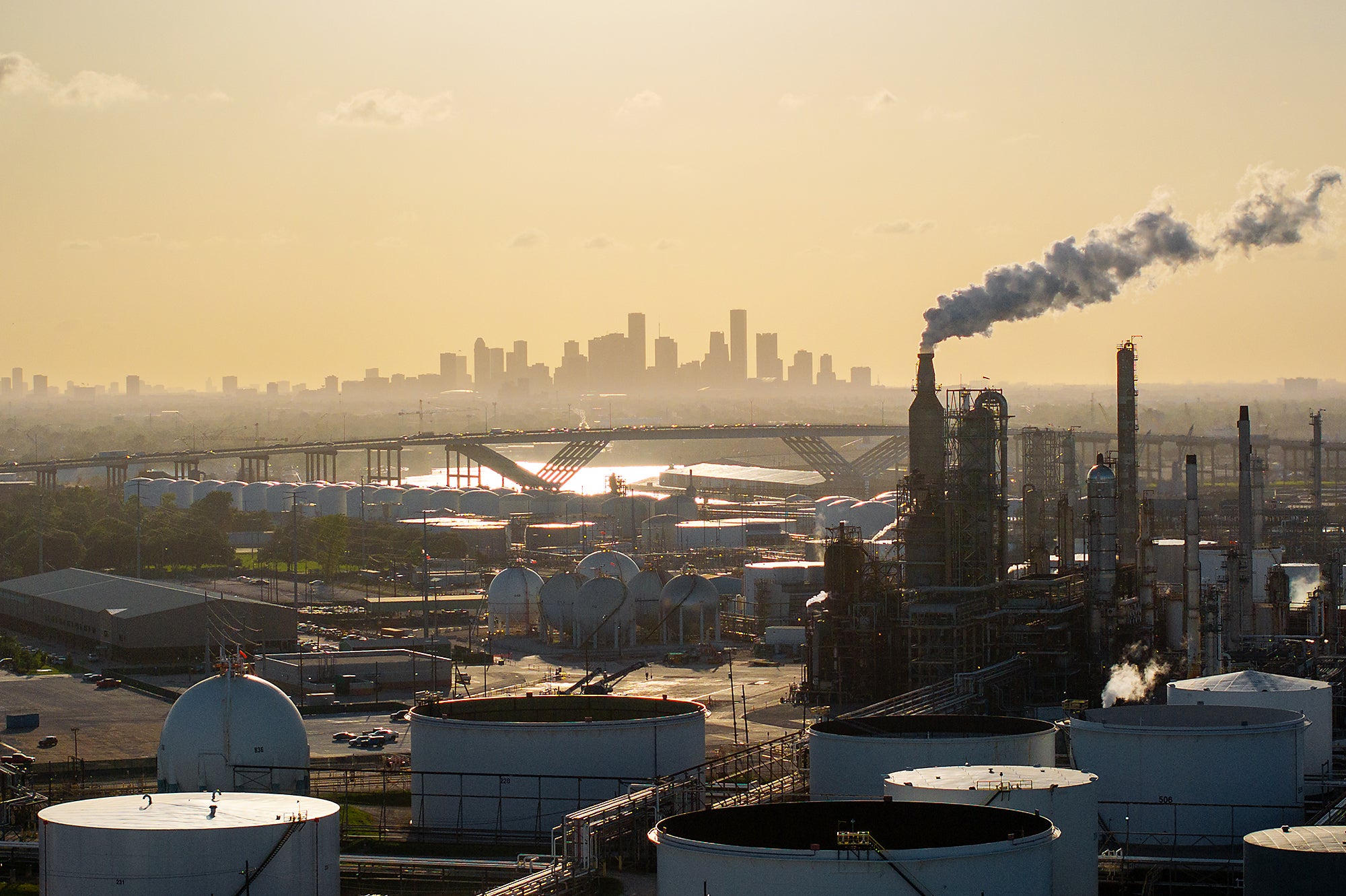 A refinery with storage tanks and exhaust stacks, some releasing vapor, are in the foreground of an aerial photo with the skyline of Houston on the horizon.