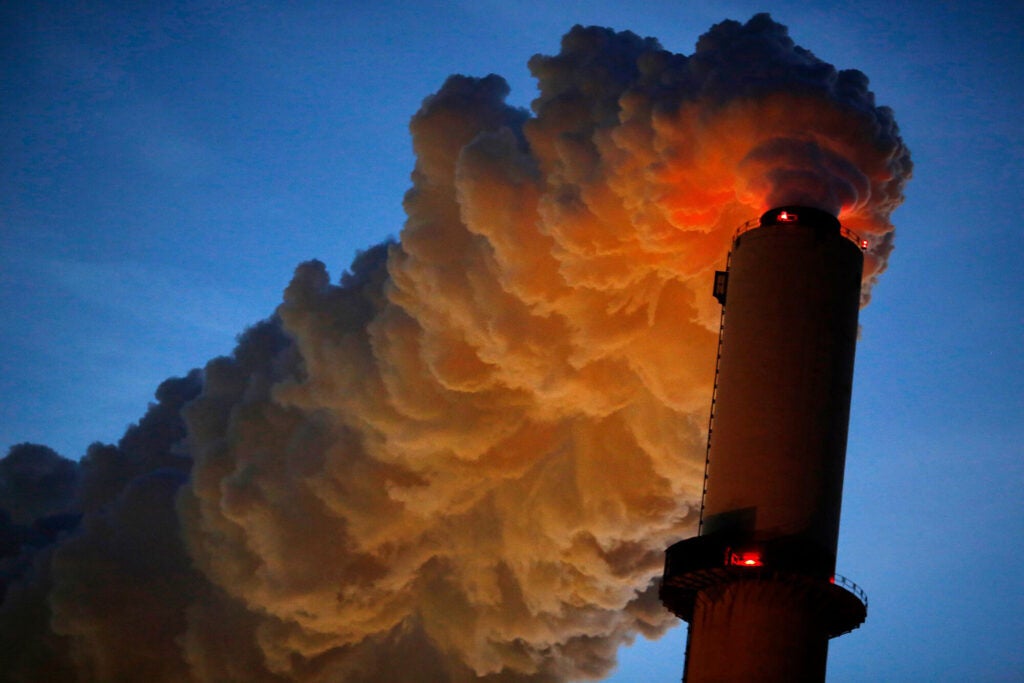 Dark gray clouds of steam emerge from the top of a smokestack at night, lit by red lights on the smokestack with a dark blue sky in the background.
