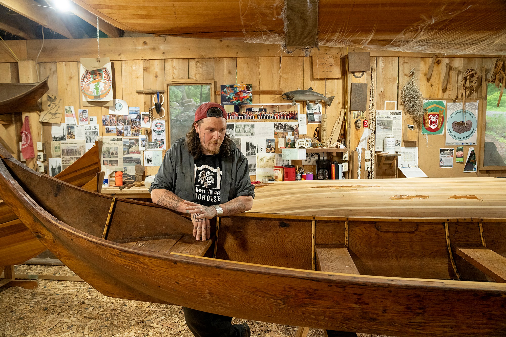 A man looks over a large wooden canoe in a wood shop with several other wooden canoes in it. On the wall behind him are tools, photographs and newspaper clippings.