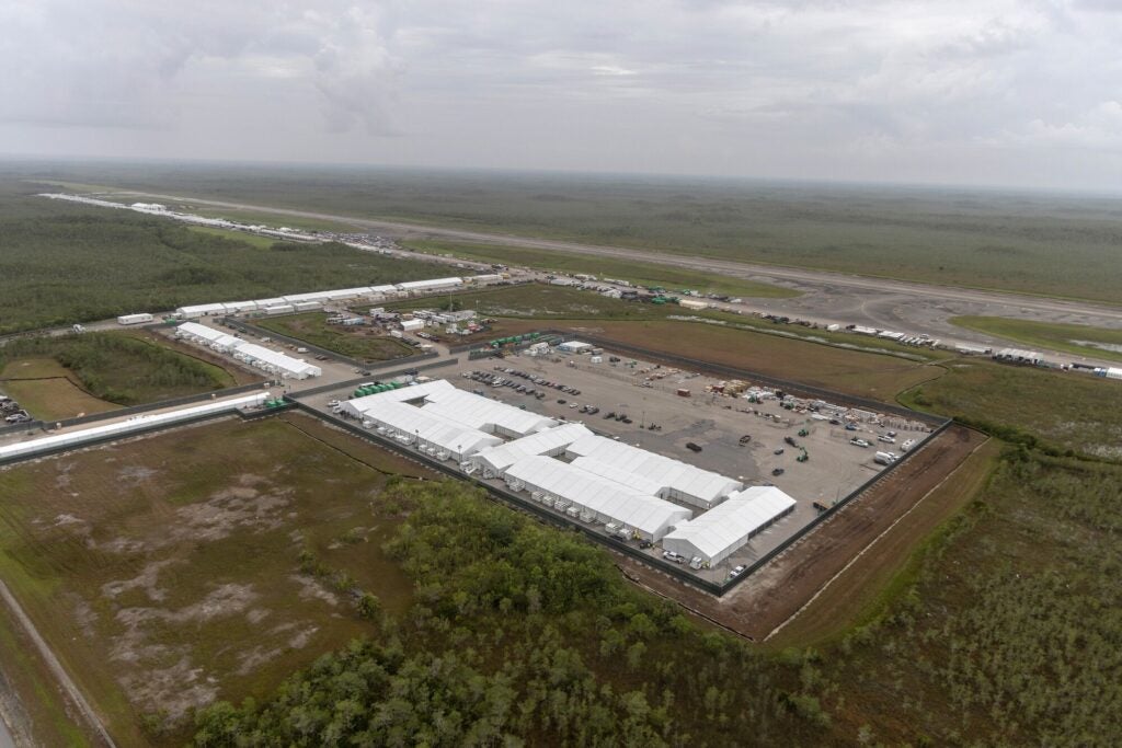 White tent buildings and vehicles fill a large parking lot next to an airstrip, surrounded by flat green land.