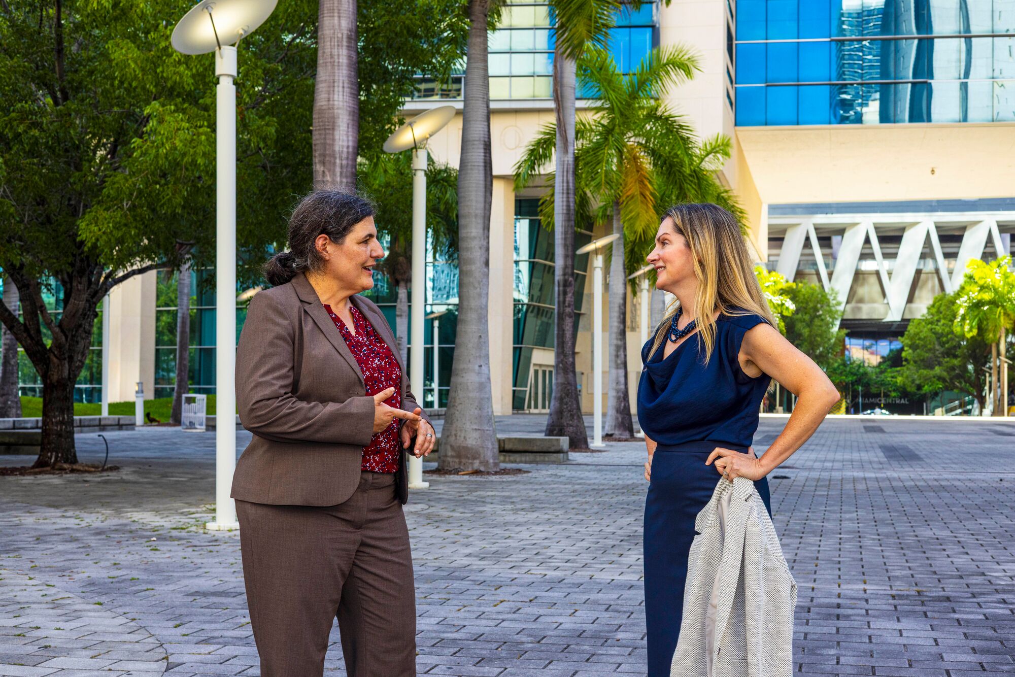 Two women stand outside of a large building surrounded by trees talking.