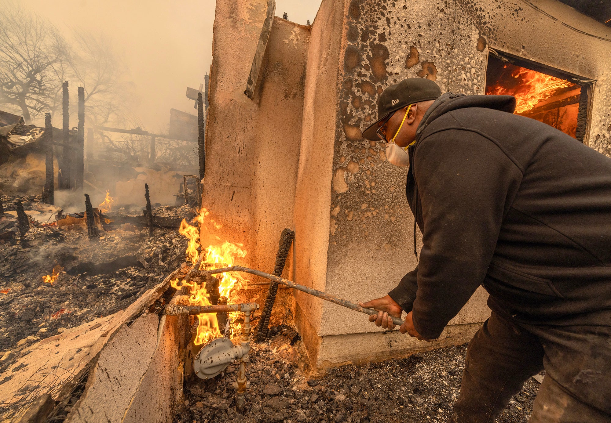 A man wearing a face mask and ball cap holds a long burned pipe stuck into a flaming set of pipes. It's next to a house which has almost completely burned and everything in the photo has also burned down, covered in smoke and small flames.