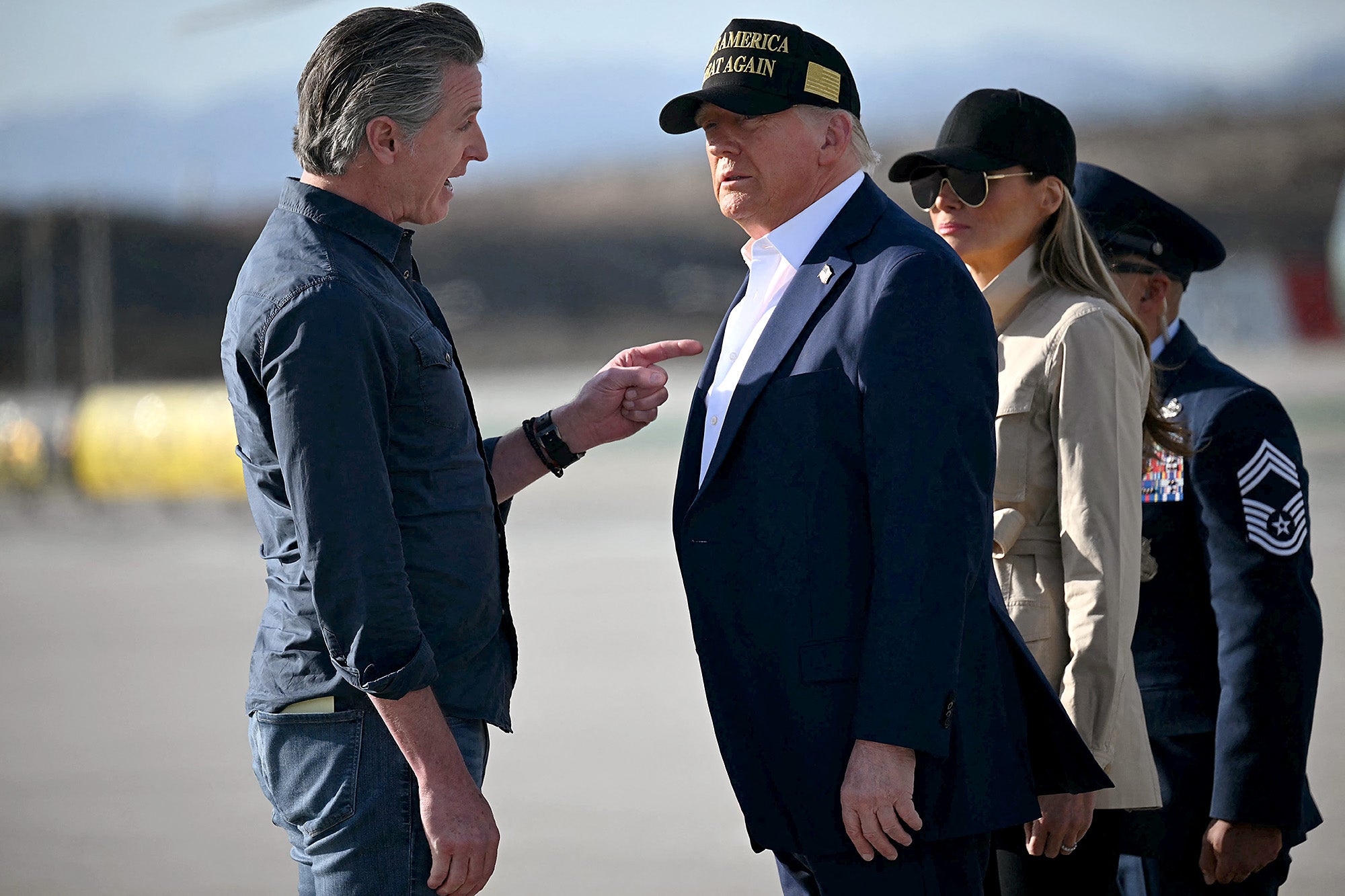 Gavin Newsom, wearing jeans and a blue collard shirt is pointing a finger at Donald Trump's chest while speaking to him with a serious expression. Trump, wearing a blue suit jacket and no tie and a black baseball hat, is looking at him.