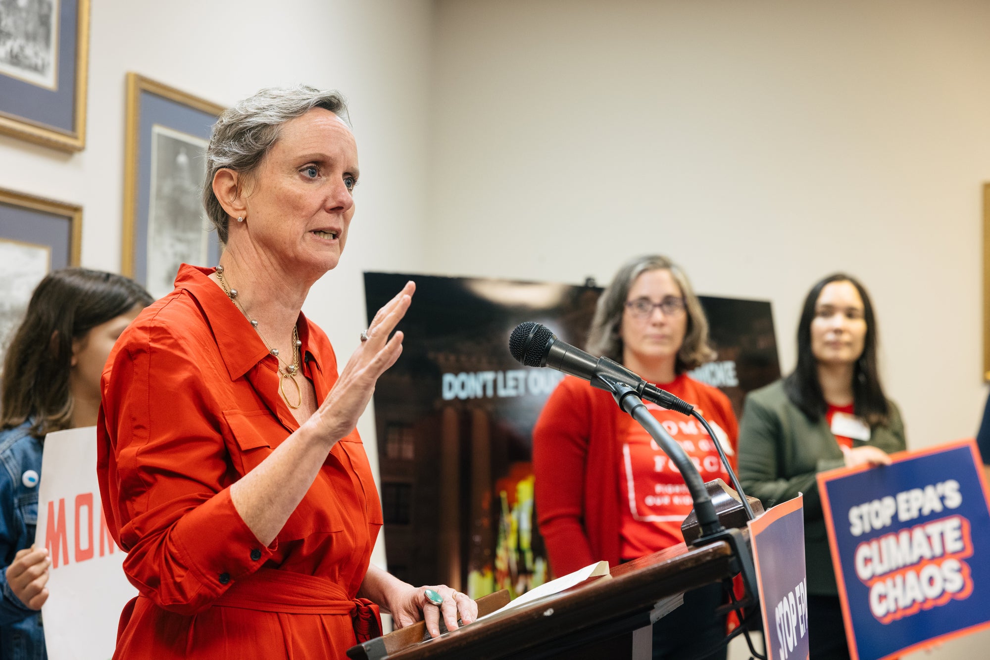 Abigail Dillen, wearing a red dress, speaks in a conference room surrounded by people holding signs that say "stop epa's climate chaos"