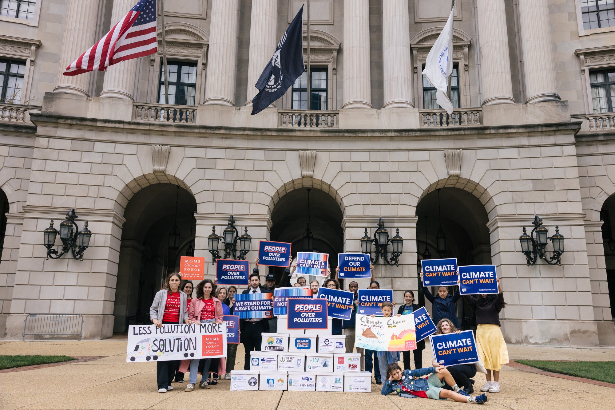 A group of people holding signs that read "climate can't wait" and "people over polluters" pose around a stack of white boxes with logos from various environmental organizations.