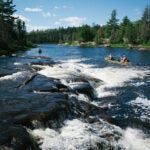 Two canoes paddle through large rapids on a wide river surrounded by green forest.