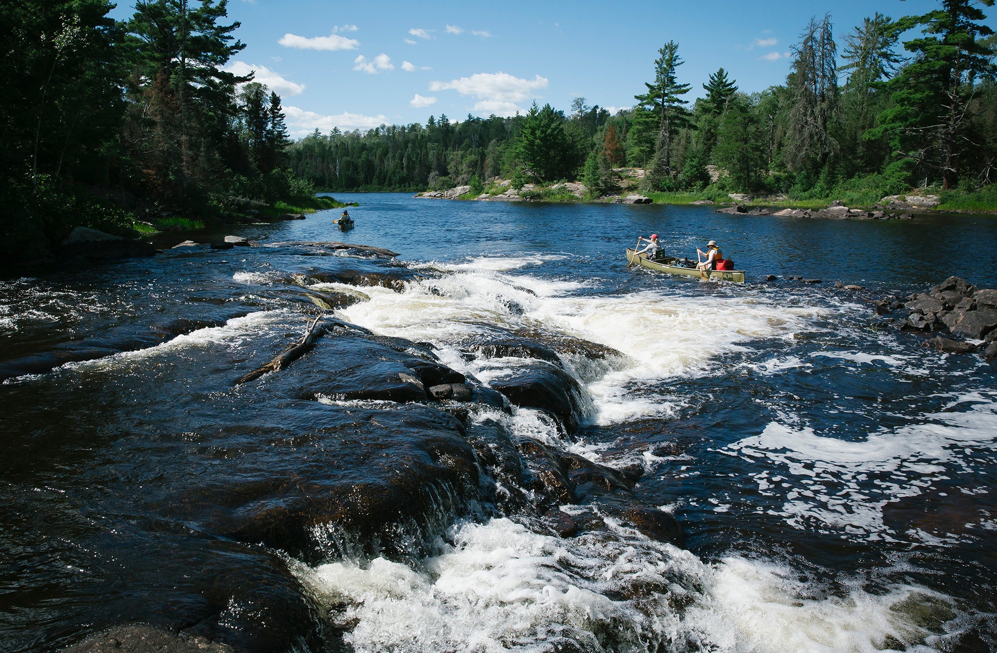 Two canoes paddle through large rapids on a wide river surrounded by green forest.