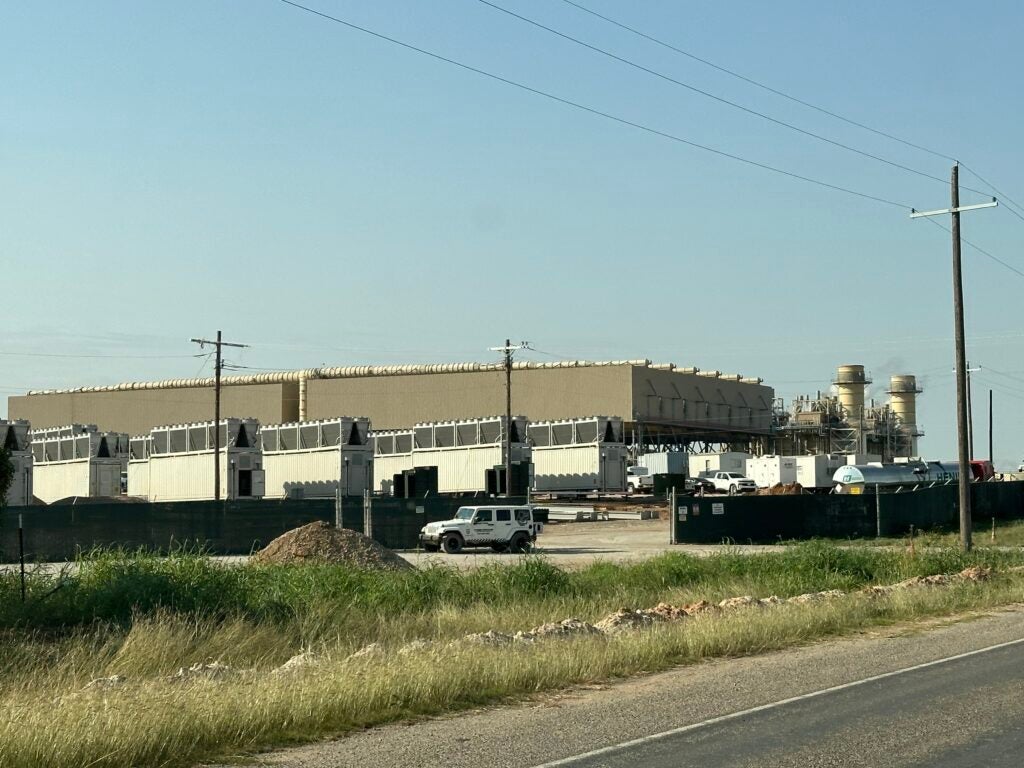 several rows of tall, white and grey cryptomining rigs and their cooling systems at MARA Cryptomine in Granbury, Texas. The Wolf Hollow gas plant is in the background, behind the rigs