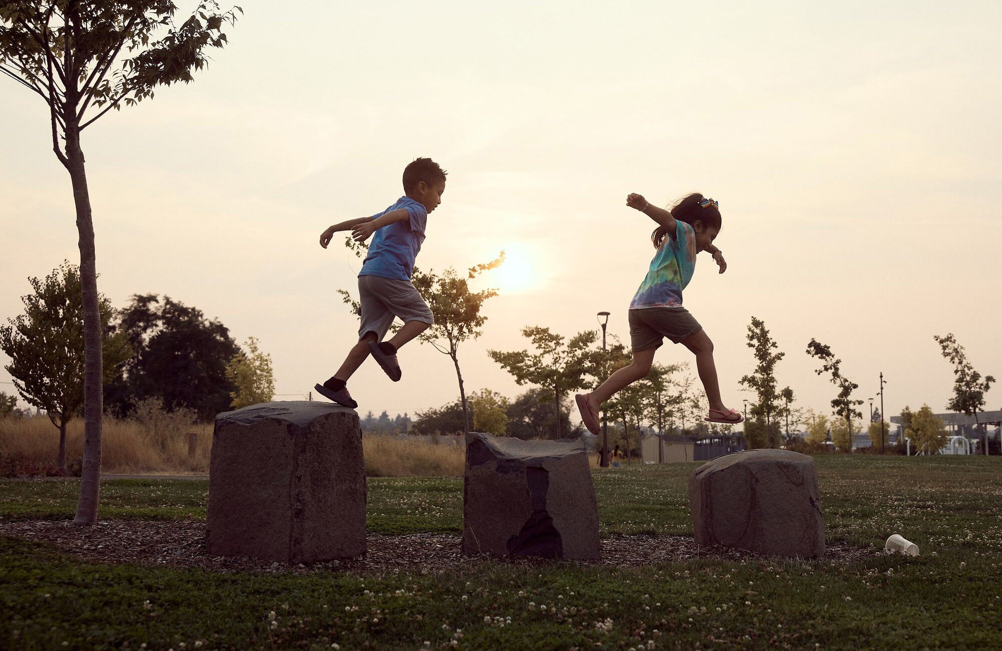Two young children leap across the tops of stone pedestals in a community park, under a hazy sky.