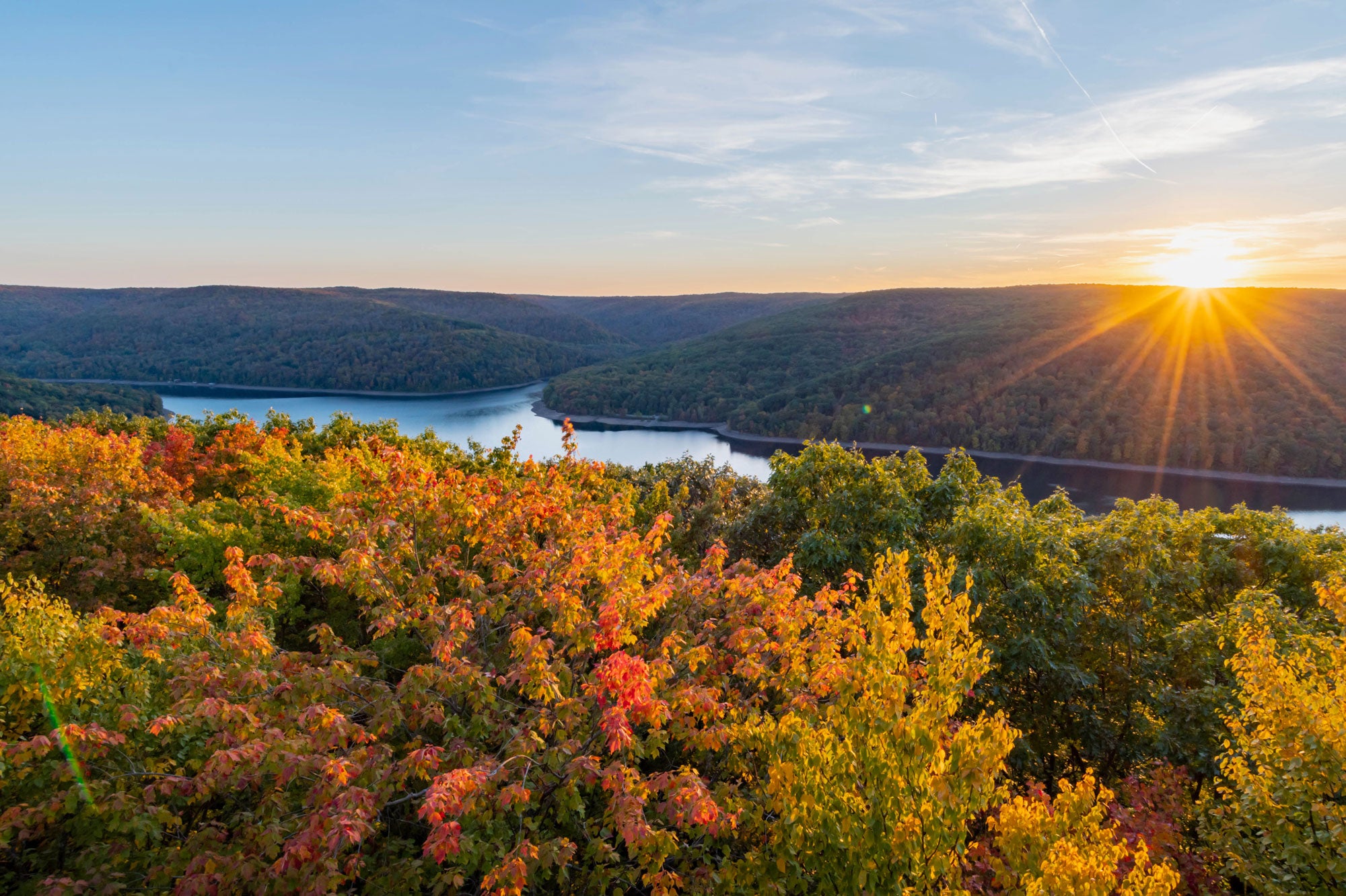 Allegheny-River_GettyImages-1599961509_2000x1333 Red, yellow, and green foliage in the foreground; a river in the middle ground; and in the background is the other bank of the river with green vegetation. The sun is just at the horizon, casting beams of rays across the background on the right side of the image.