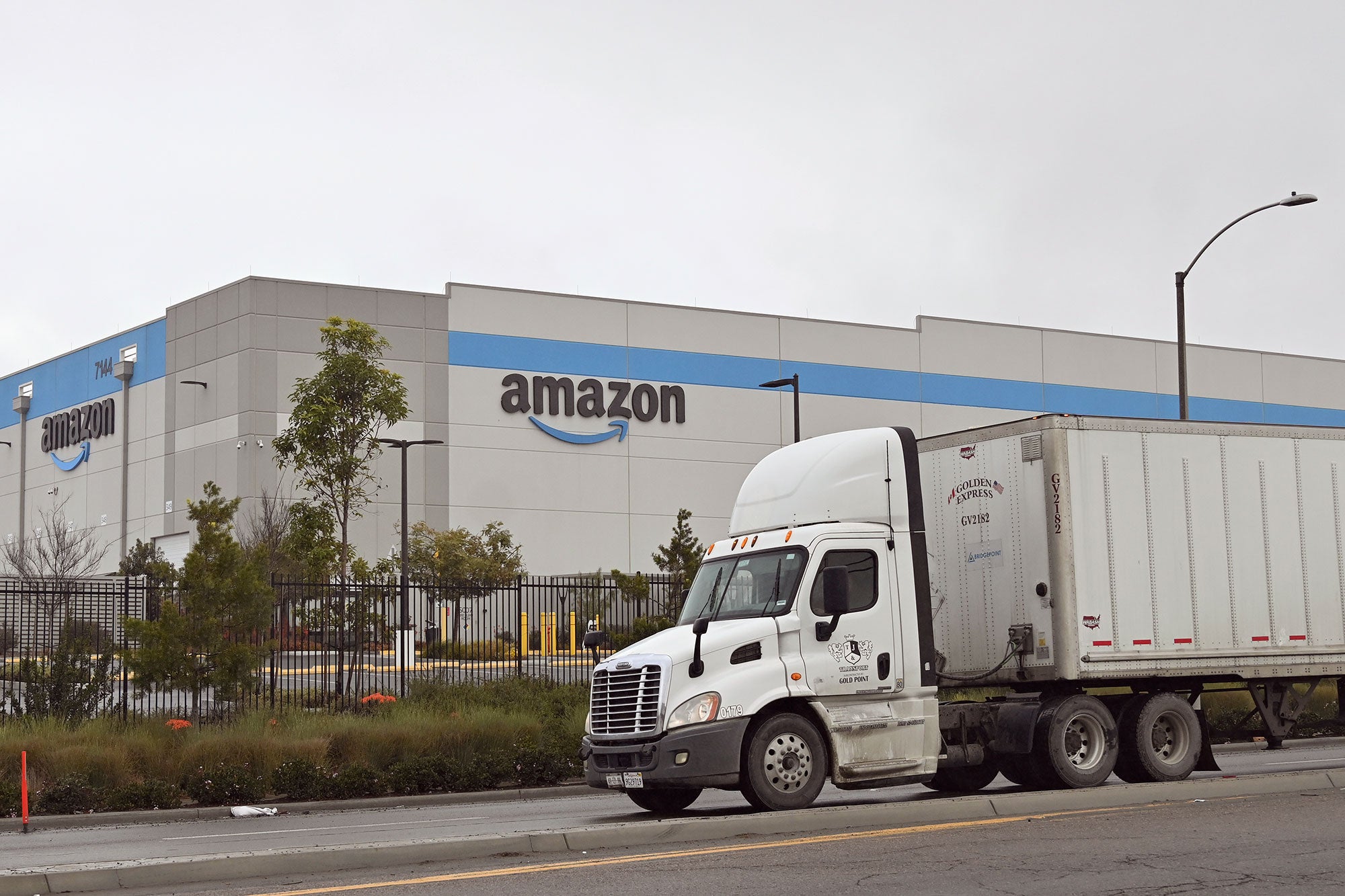 A white trailer truck is stopped on the street outside of a large Amazon warehouse on an overcast day.