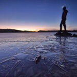 A backlit person stands on a rocky outcropping, surrounded by icy water and framed against the twilight in the Boundary Waters Canoe Area Wilderness.