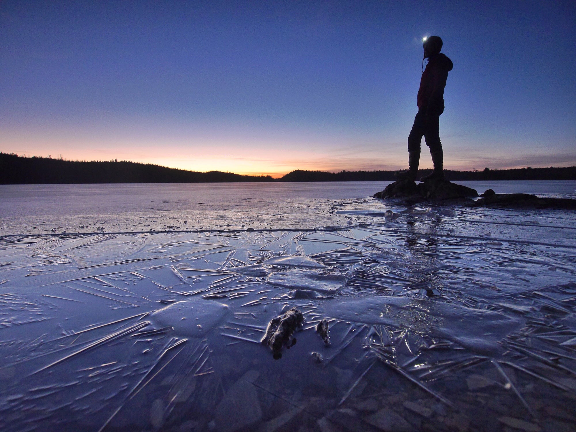 A backlit person stands on a rocky outcropping, surrounded by icy water and framed against the twilight in the Boundary Waters Canoe Area Wilderness.