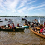 Many people in kayaks float near the shore in the Boundary Waters Canoe Area Wilderness. Some hold signs opposing the Twin Metals mine.