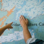Two people in front of a map of the Boundary Waters Canoe Area Wilderness. One points to a location on the map. while the other holds an open hand against the border line with Canada.