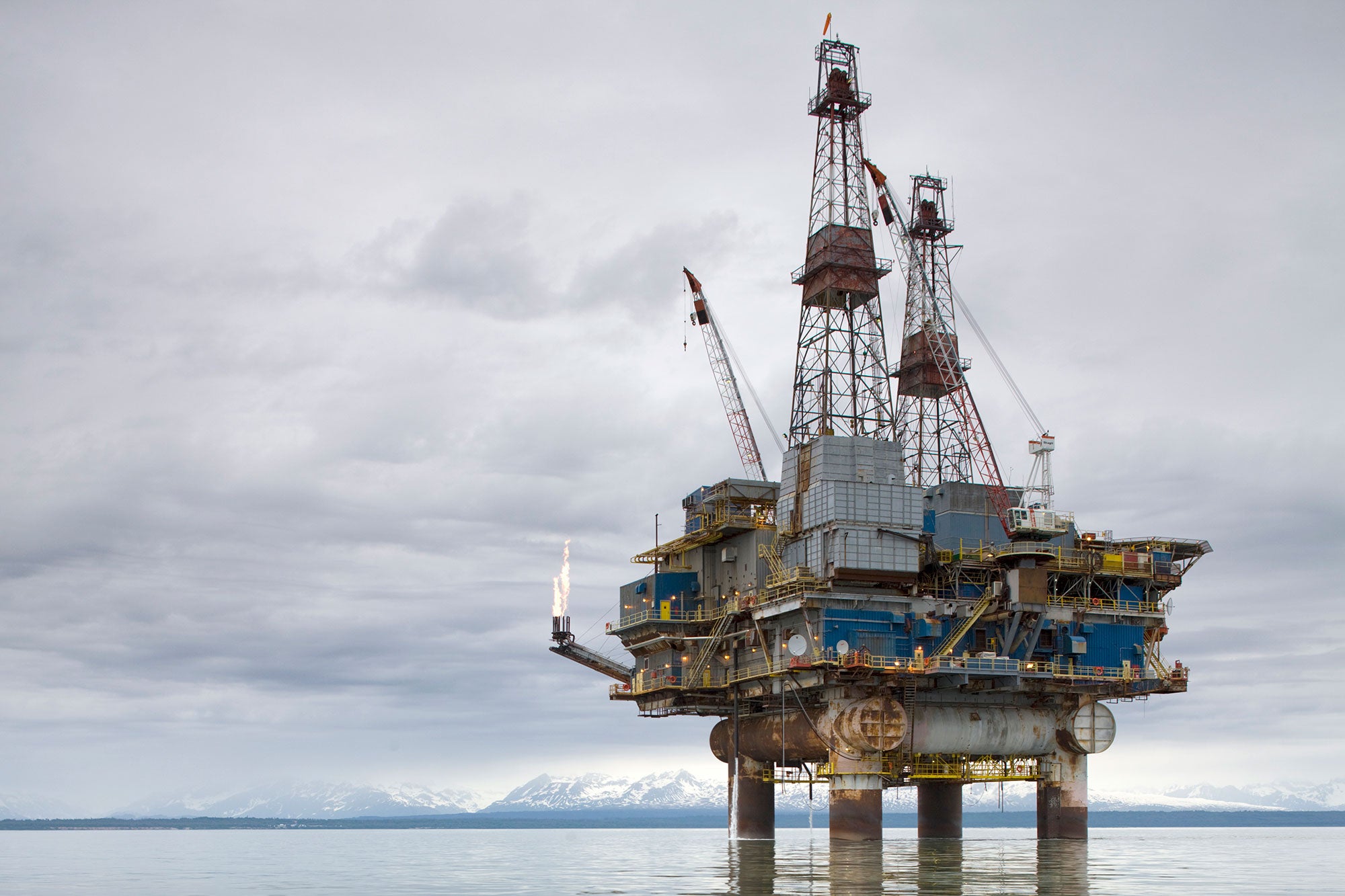 A rusty offshore oil drilling platform stands in the ocean, with a large flare burning from one corner. Snow-capped mountains are in the background.