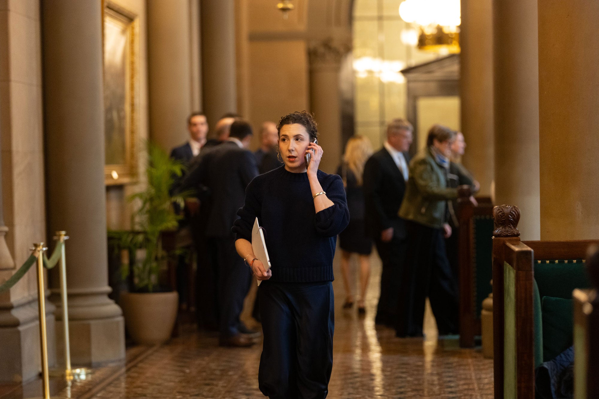 ElectifyNY-Advocacy_elizabeth-moran_2000x1333 A woman in a navy sweater and navy skirt is walking, with documents tucked against her right side with her right arm. With her left hand, she is holding a mobile phone to her ear. A background of blurred people in suits.