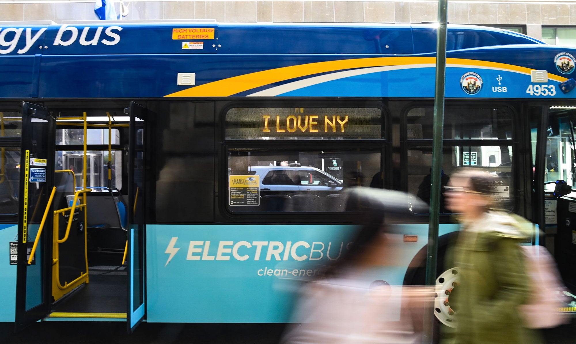 Electric-Bus---NYC_2000x1193 The side of a blue electric bus with the doors open. A display above the window reads "I Love NY." Blurred pedestrians in motion in front of the bus.