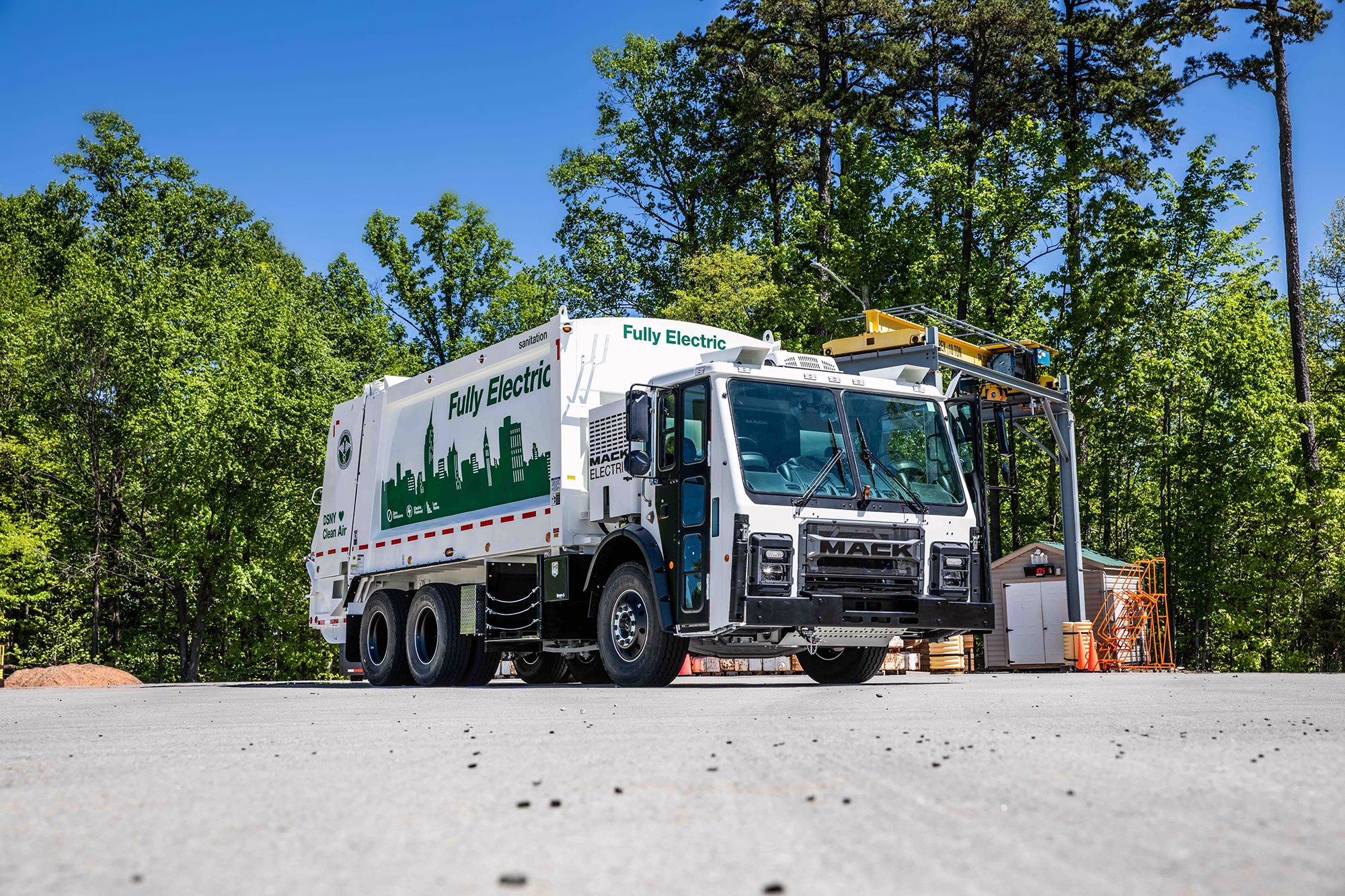A white Mack garbage truck with the words "Fully Electric" and "Clean Air" painted on its side is parked in an empty open lot next to a small shed, surrounded by bright green trees.