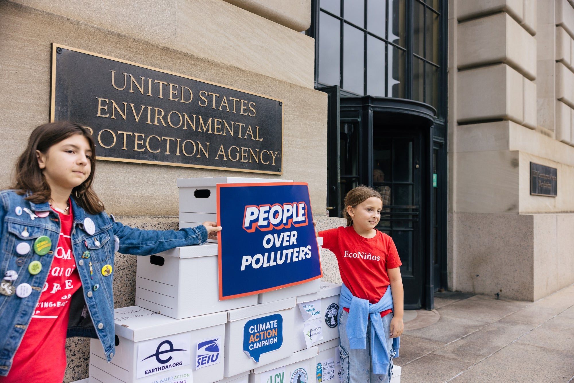 Two youngsters wearing bright red T-shirts -- one shirt says "EcoNinos" -- hold a sign that reads "People Over Polluters" in front of a large tower of boxes containing public comment letters at the entrance to the U.S. Environmental Protection Agency.