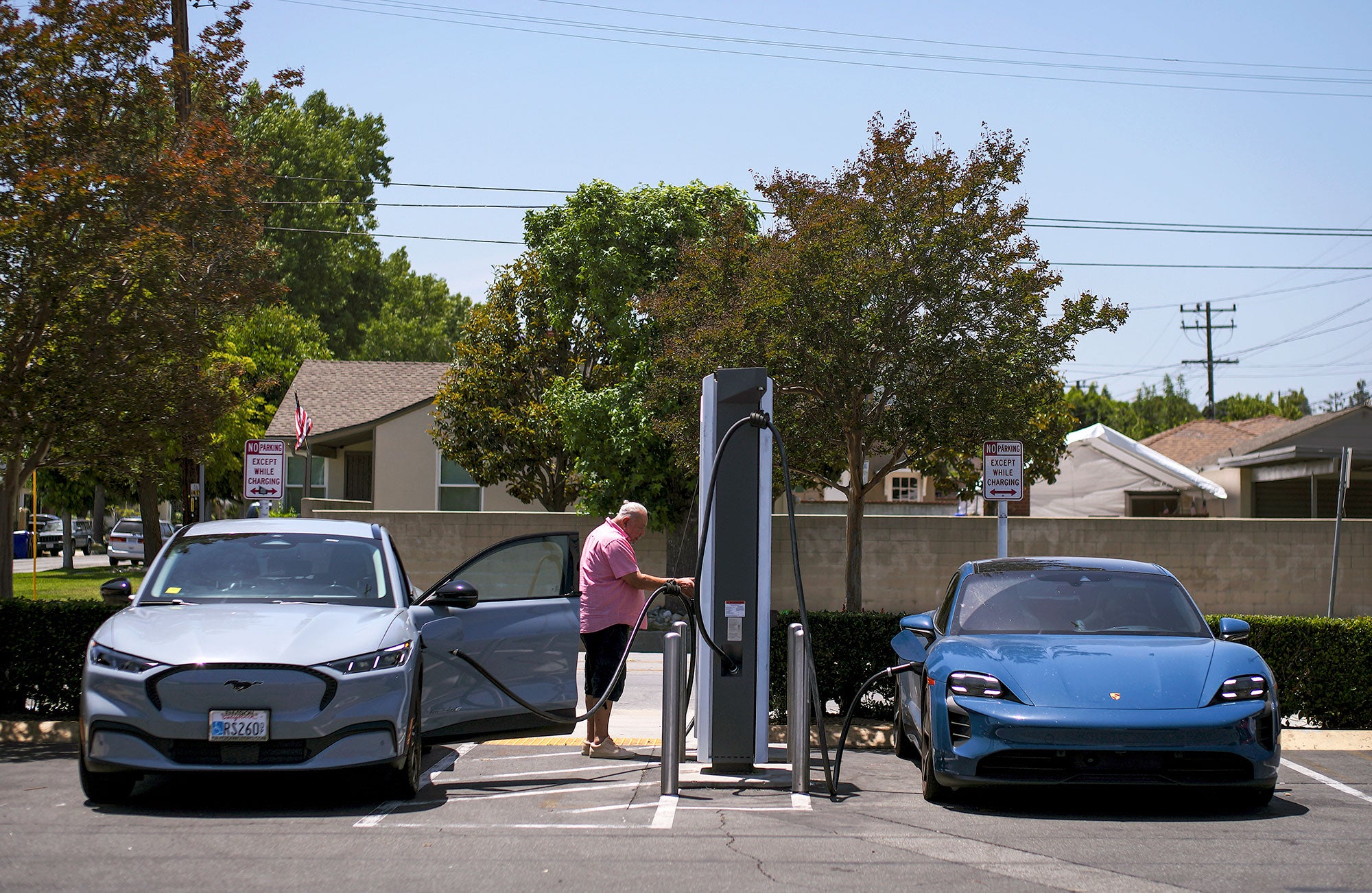 Two electric passenger cars -- one blue-grey and the other bright blue -- are parked and plugged into an electric charging station in a parking lot surrounded by trees and homes. A person in a pink shirt stands by an opened door of the blue-grey electric car at the charging station.