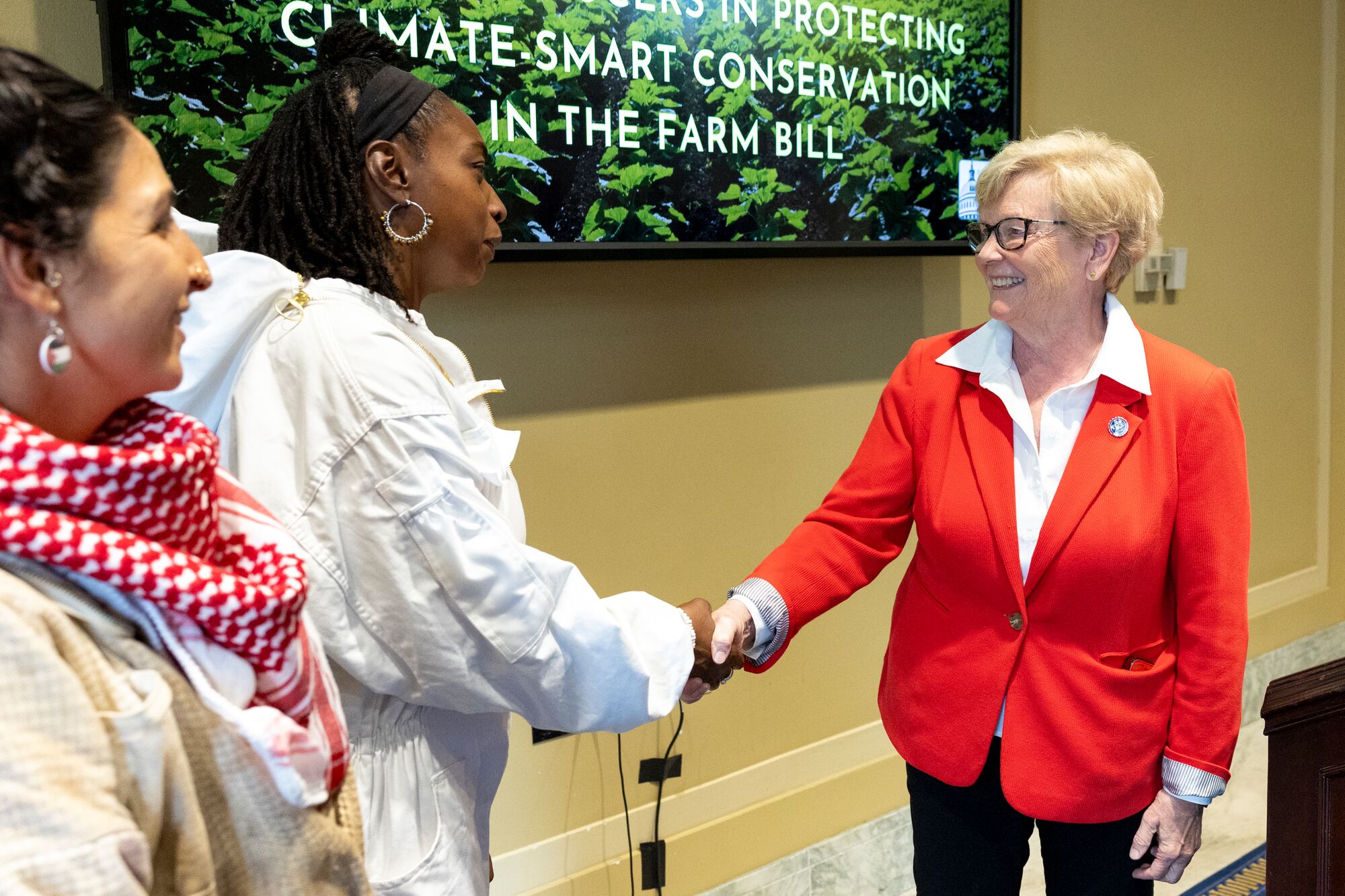 Farm Bill 2024 Lobby Day on Capitol Hill A person wearing a protective beekeeping suit speaks and shakes hands with a smiling person wearing a bright red suit jacket in front of a large TV screen displaying the text "Climate-smart conversation in the Farm Bill."