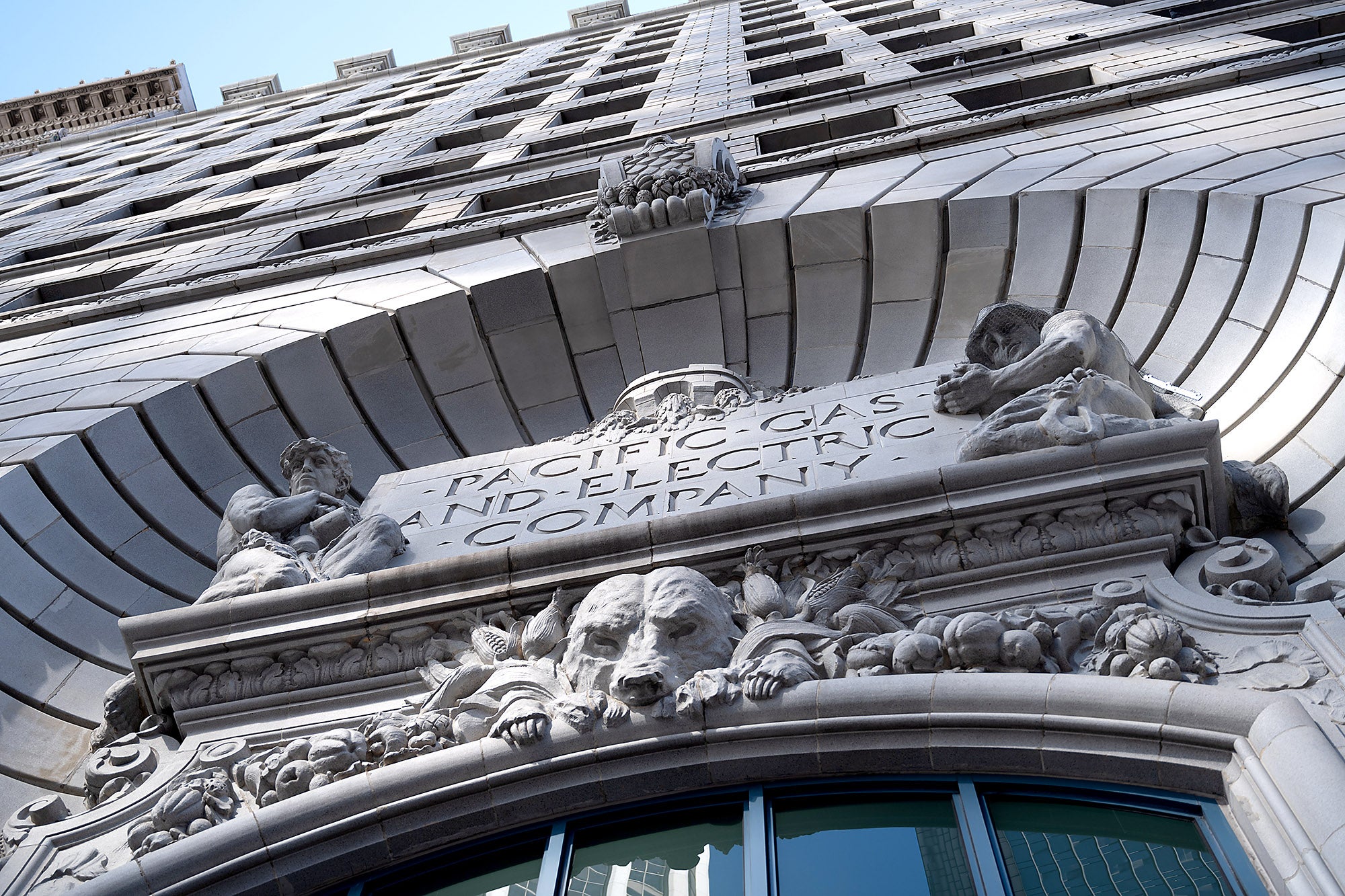 The front door of a stone office building built in the 1920. The photo is looking up, only seeing the ornate carved stonework above the door, which includes a sign saying "Pacific Gas and electric Company. The sign is flanked by the statues of two men in an ancient Greek or Roman style.