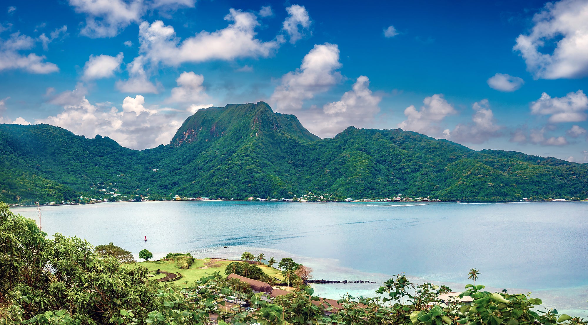 Pago Pago Harbor on Tutuila Island A wide landscape view of a green island wrapping around a large, natural, blue ocean harbor. Small buildings are on the coast with a tall hill or mountain on the far side.