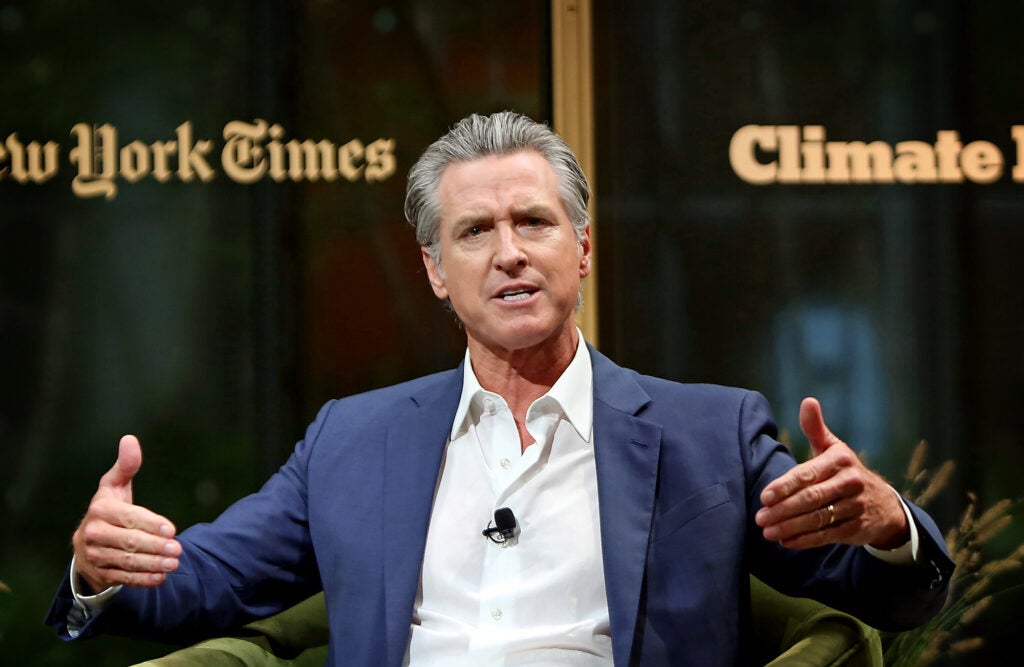 Gavin Newsom, wearing a blue sports jacket and white shirt gestures with both hands while speaking.