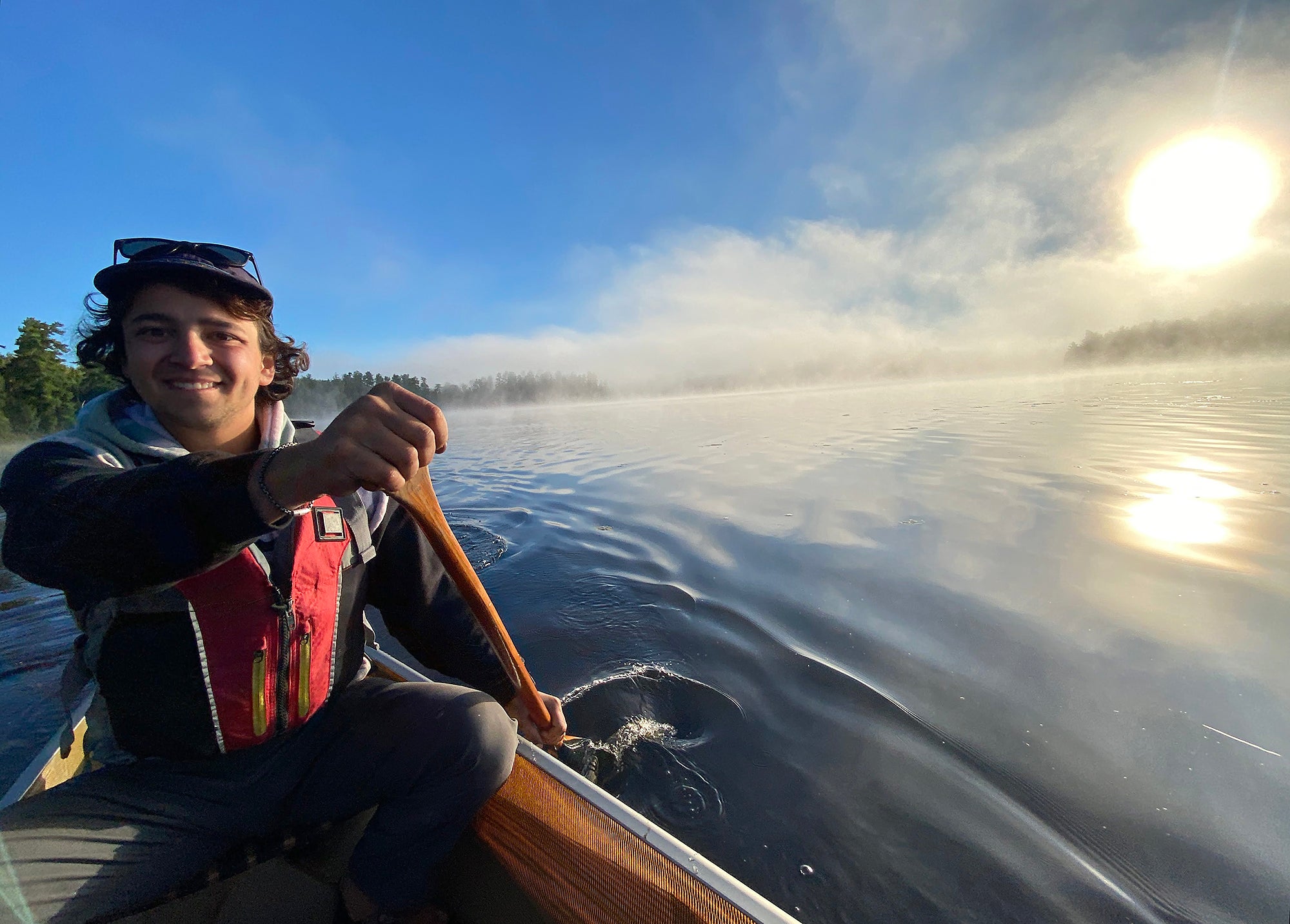 A young man in a life preserver sitting in a canoe smiles and looks into the camera while paddling in a misty lake with a low sun.