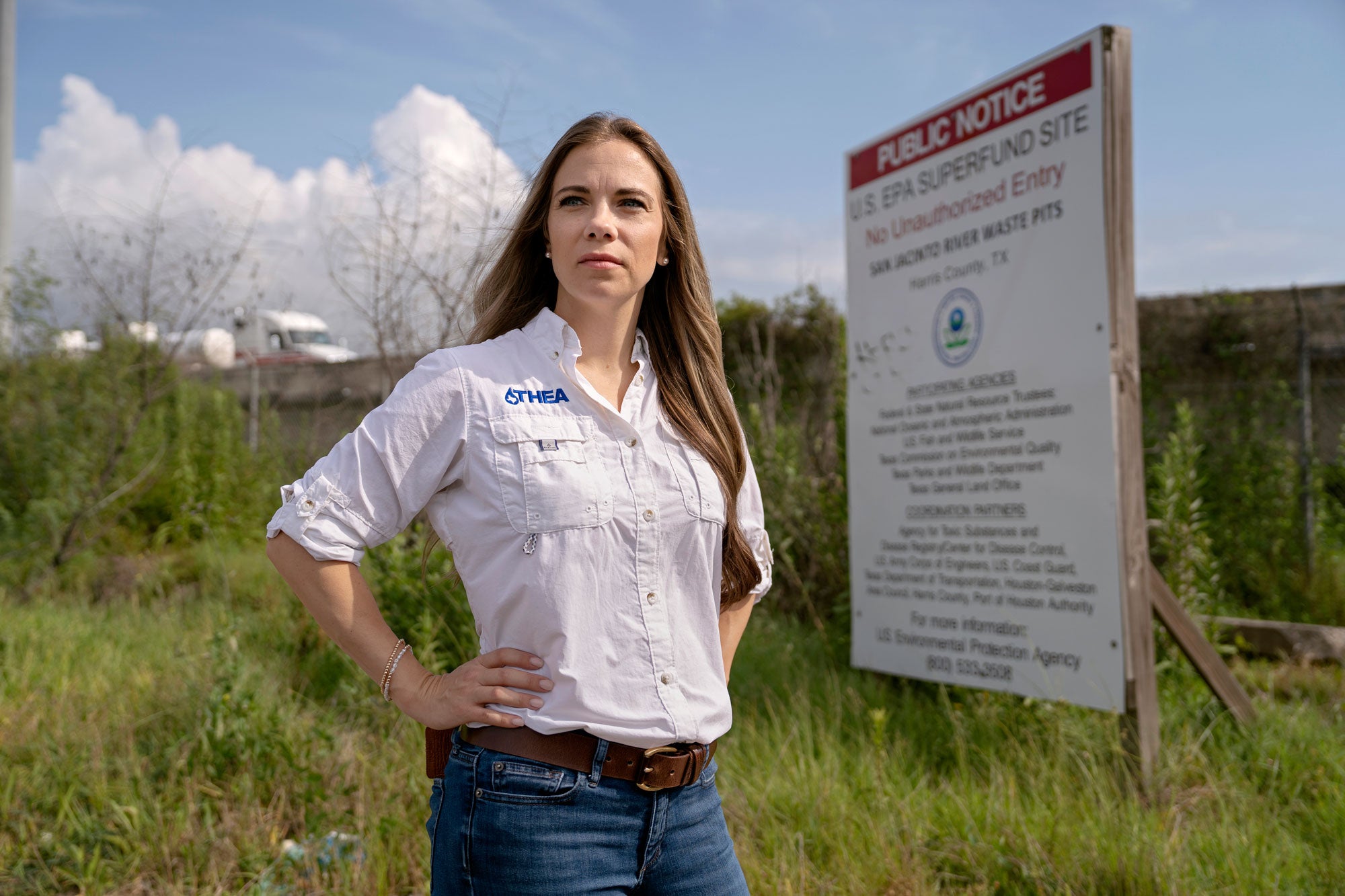 A woman stands in front of a public notice of an U.S. EPA Superfund Site sign. She is wearing a light-colored button-up shirt with THEA embroidered above a chest pocket.