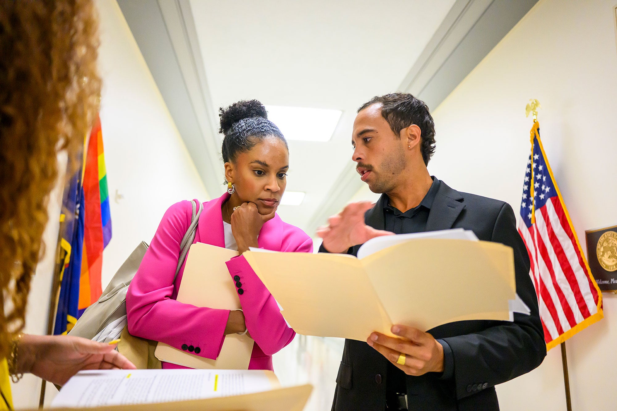 Two people stand talking in a brightly lit hallway, while one holds an open manila folder.