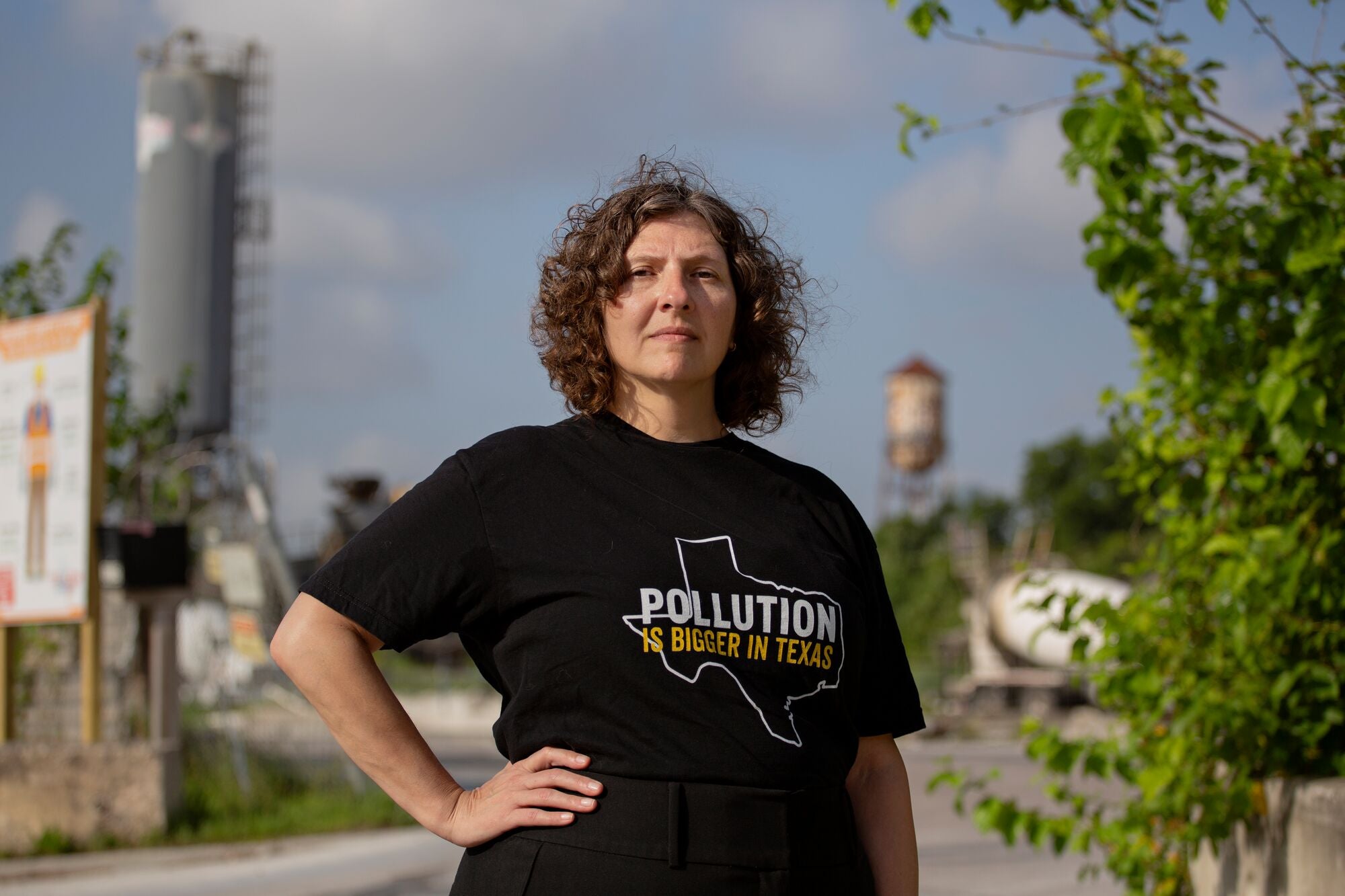 A person wearing a black shirt that reads "Pollution is bigger than Texas" stands in front of an industrial facility under a grey-blue sky.