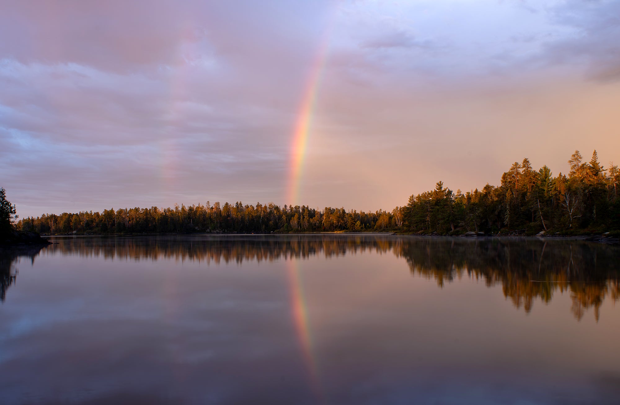 A wide landscape photo with a calm lake in the foreground, a line of trees midway and a lightly cloudy sky above. A rainbow, reflected in the lake, cuts across the center of the photo.