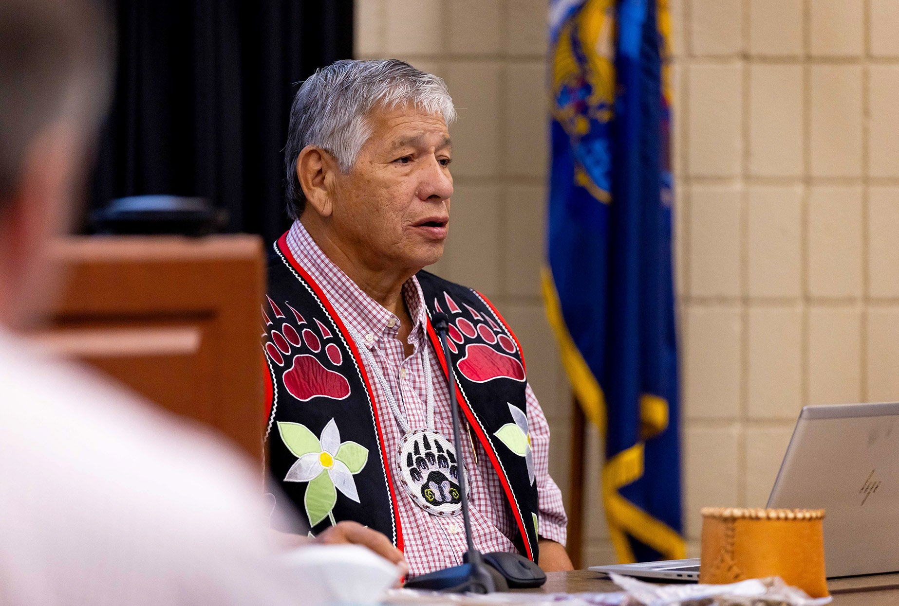 A man with a black and red vest decorated with a pattern of bear claws and flowers speaks into a microphone while seated at a table.