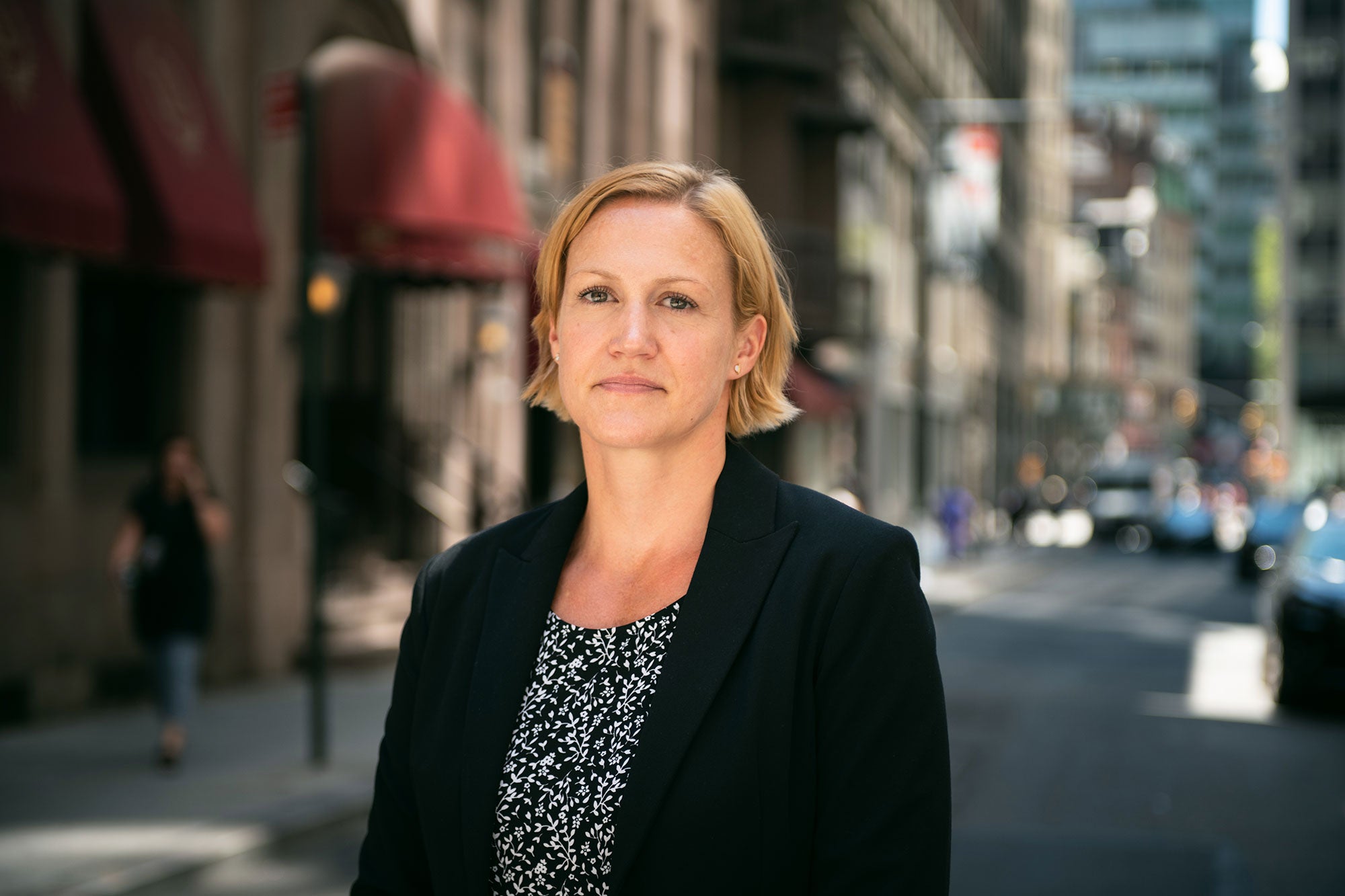 Mandy DeRoche, wearing a black suit jacket over a black blouse with a white flower pattern, stands on a busy New York City street.