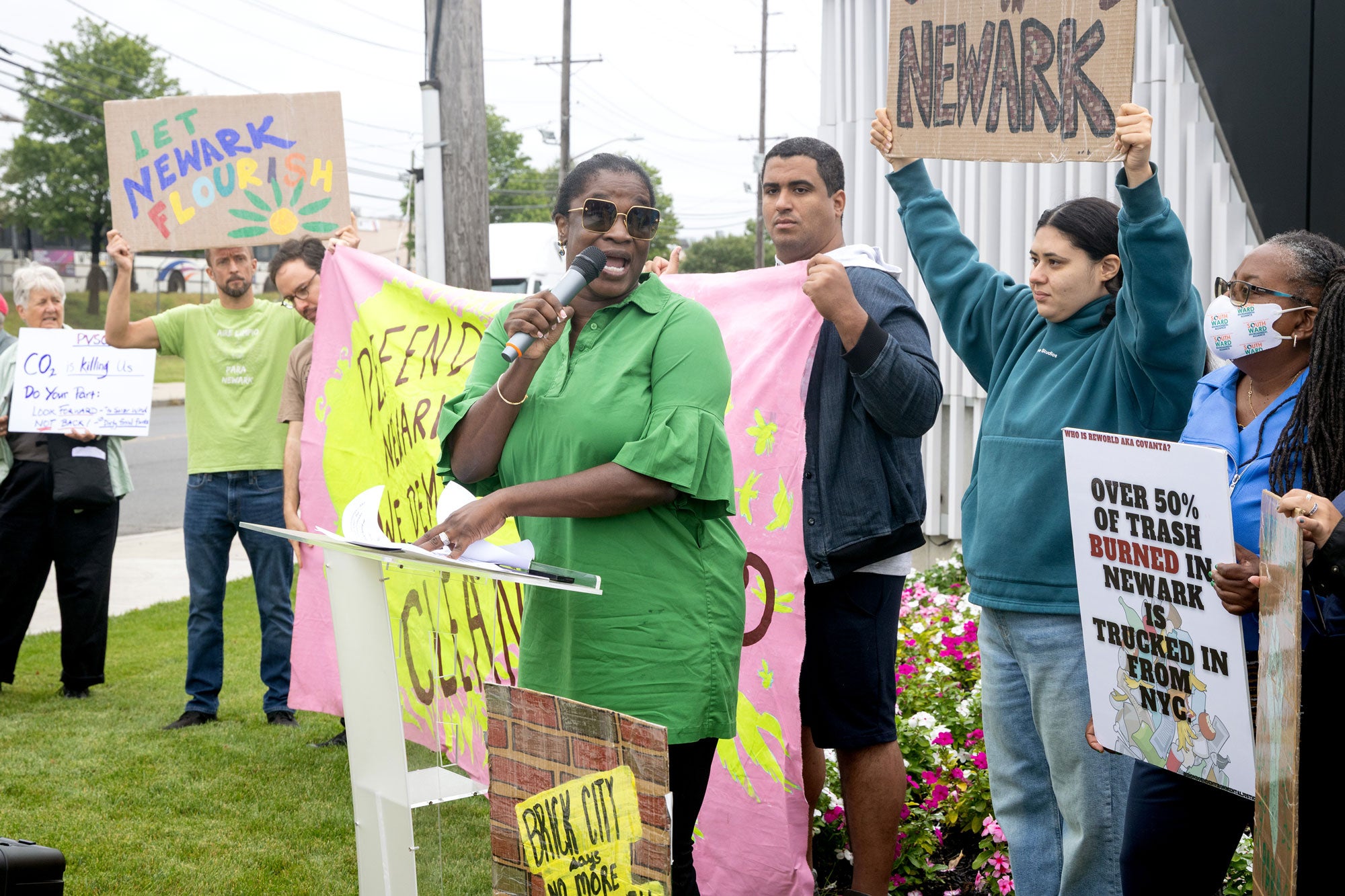 Newark-Environmental-Justice-Press-Conference-and-Pollution-Tour_2000x1333 Protestors holding signs that say "Let Newark Flourish," "Over 50% of trash burned in Newark is trucked in from NYC," and more. A woman dressed in bright green and sunglasses, stands in front of a podium with a mic in hand, in front of the crowd of protestors.
