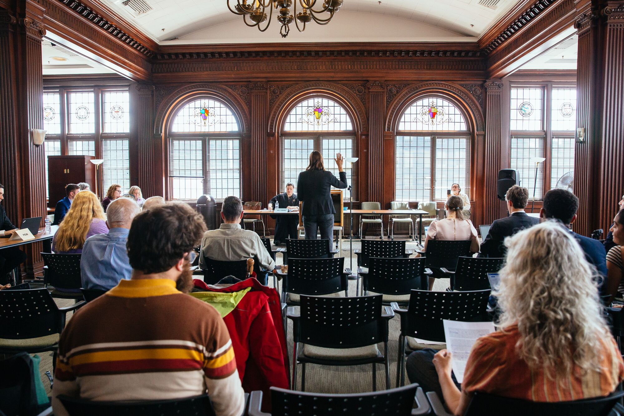In a large light-filled hall with a wall of stained glass windows, a person stands with their right hand raised before a seated person in black robes. Behind the standing person are rows of chairs, half-filled with seated people, some of whom are looking through papers.