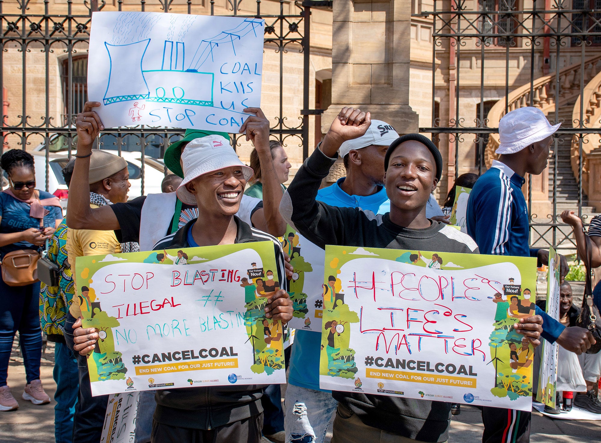 A crowd of passionate people stand outside the black wrought iron fence of an ornate building on a sunny day, holding signs that read, "Coal Kills Us. Stop Coal," "Stop Illegal Blasting: No More Blasting," and "#People's Life's Matter."