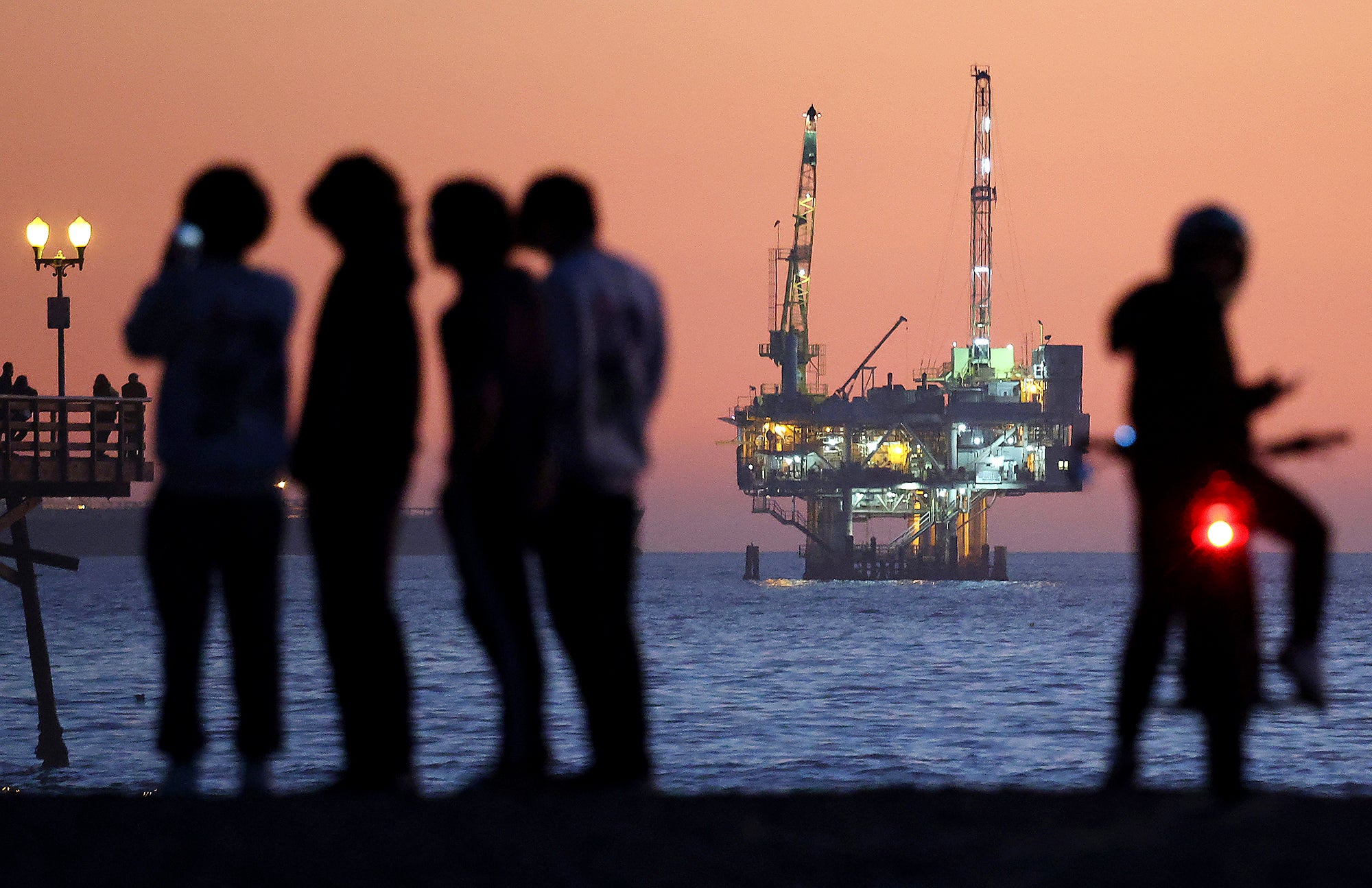 A group of silhouetted people stand on the shore in the evening with a lit up drilling platform visible in the distance.